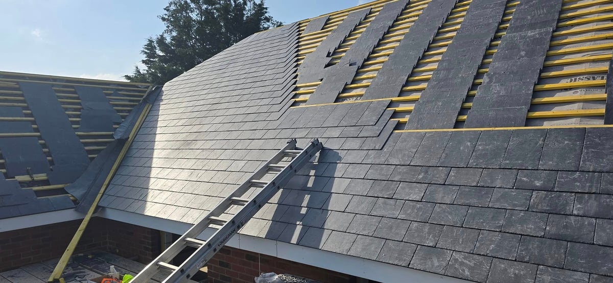 Roof under construction with shingles and yellow wooden trusses, metal ladder leaning against the slope