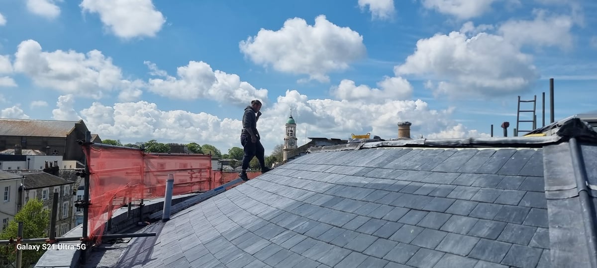 Worker on gray slate roof with red container and industrial background under blue sky with clouds