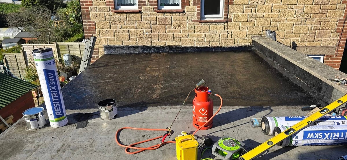 Flat roof waterproofing in progress with worker in orange clothing and hose beside brick building